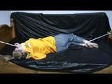 Samantha in a shiny yellow rain jacket and a shiny grey rain trowsers tied and gagged on bed (Video)