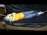 Samantha in a shiny yellow rain jacket and a shiny grey rain trowsers tied and gagged on bed (Video)