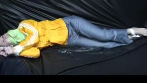 Samantha in a shiny yellow rain jacket and a shiny grey rain trowsers tied and gagged on bed (Video)