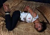 Barefoot Christy in white t-shirt and black jeans, hogtied with hemp rope, wiggles on the bed