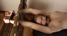Ballgagged barefoot topless Albina Rose, lying on the table with her feet tied under the table and with her cuffed hands locked in the stocks