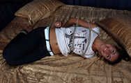 Barefoot Christy in white t-shirt and black jeans, hogtied with hemp rope, wiggles on the bed