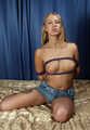 Topless barefoot Nancy in jeans shorts, hogtied with her hands in reverse prayer, wiggles on the bed
