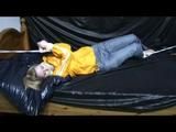 Samantha in a shiny yellow rain jacket and a shiny grey rain trowsers tied and gagged on bed (Video)