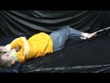 Samantha in a shiny yellow rain jacket and a shiny grey rain trowsers tied and gagged on bed (Video)