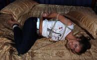 Barefoot Christy in white t-shirt and black jeans, hogtied with hemp rope, wiggles on the bed