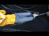 Samantha in a shiny yellow rain jacket and a shiny grey rain trowsers tied and gagged on bed (Video)