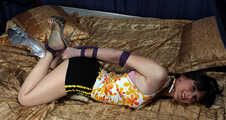 Karen in yellow top, black shorts and mules, hogtied with her feet crossed and her elbows tightly bound, wiggles on the bed