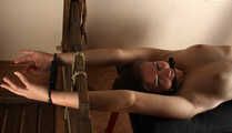 Ballgagged barefoot topless Albina Rose, lying on the table with her feet tied under the table and with her cuffed hands locked in the stocks