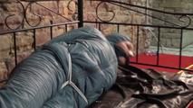 Mara tied, gagged and hooded on bed in a cellar wearing a shiny blue/grey downwear (Video)