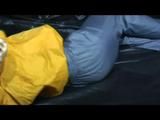 Samantha in a shiny yellow rain jacket and a shiny grey rain trowsers tied and gagged on bed (Video)
