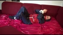 Lucy tied and gagged with ropes and a cloth gag on a sofa wearing a blue/red rainwear combination (Video)