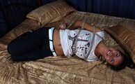 Barefoot Christy in white t-shirt and black jeans, hogtied with hemp rope, wiggles on the bed