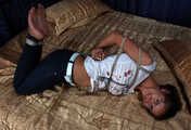 Barefoot Christy in white t-shirt and black jeans, hogtied with hemp rope, wiggles on the bed