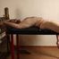 Ballgagged barefoot topless Albina Rose, lying on the table with her feet tied under the table and with her cuffed hands locked in the stocks