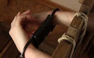 Ballgagged barefoot topless Albina Rose, lying on the table with her feet tied under the table and with her cuffed hands locked in the stocks