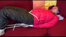 Mara tied and gagged on a sofa wearing a shiny pink down jacket and a black rain pants (Video)