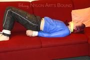 Mara tied and gagged on a sofa wearing a shiny black rain pants and a sexy lightblue down jacket (Video) 