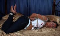Barefoot Christy in white t-shirt and black jeans, hogtied with hemp rope, wiggles on the bed