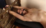 Ballgagged barefoot topless Albina Rose, lying on the table with her feet tied under the table and with her cuffed hands locked in the stocks