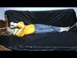 Samantha in a shiny yellow rain jacket and a shiny grey rain trowsers tied and gagged on bed (Video)
